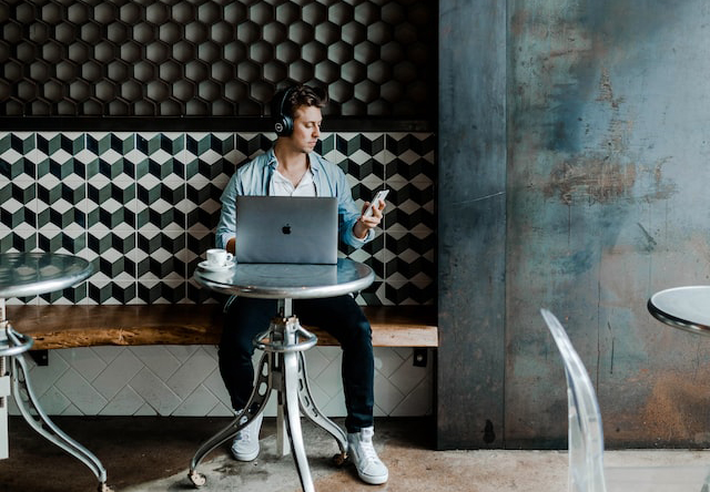 Junger Mann mit Laptop am Tisch und Handy in der Hand in einem Lokal