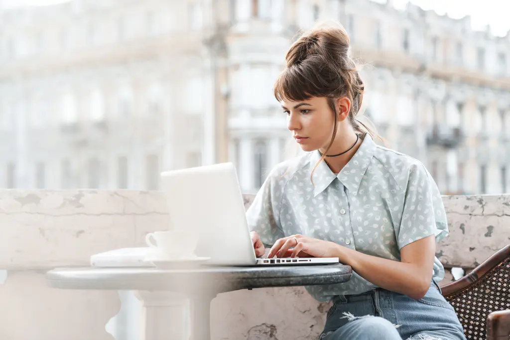 Eine Person mit geschlossenen, dunklen Haaren sitzt auf einem Balkon an einem Tisch und blickt auf den Bildschirm eines aufgeklappten Laptops. Daneben sind eine Tasse und ein Buch. Hinter der Person ist ein Gebäude